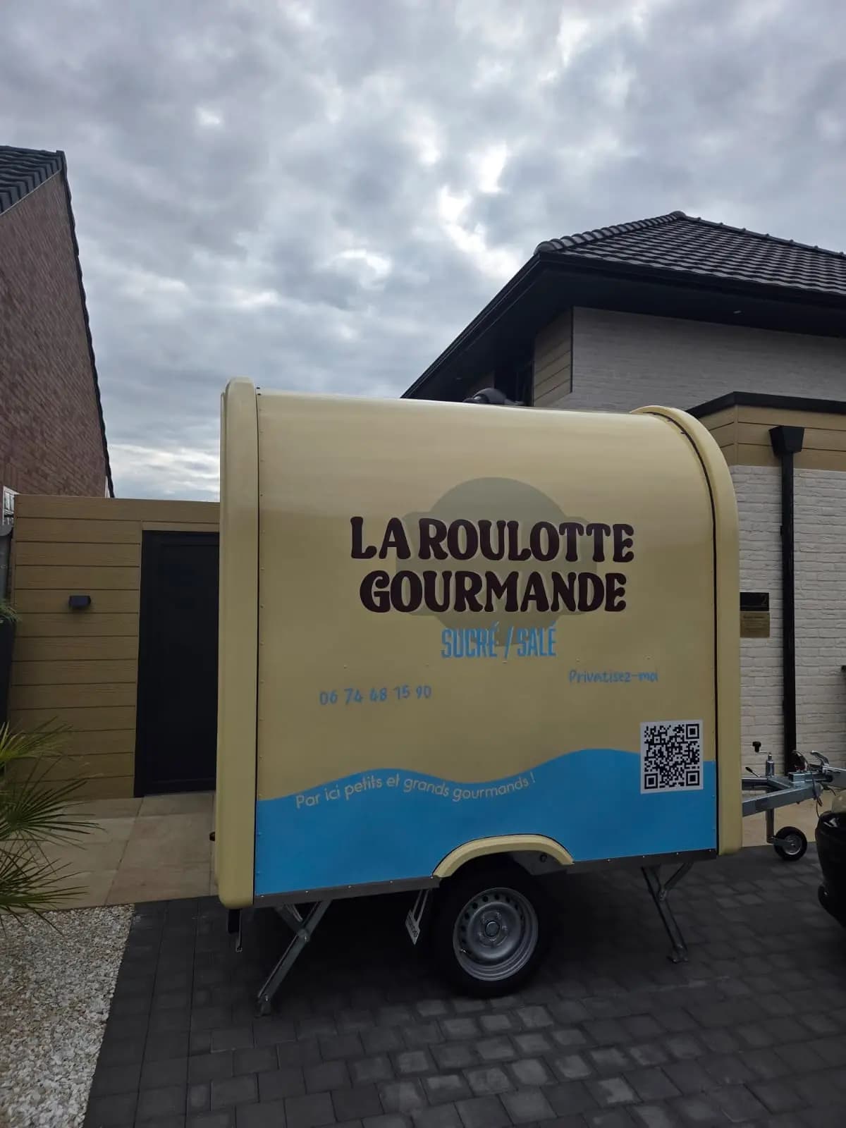 Remorque food truck La Roulotte Gourmande beige et bleue stationnée devant une maison